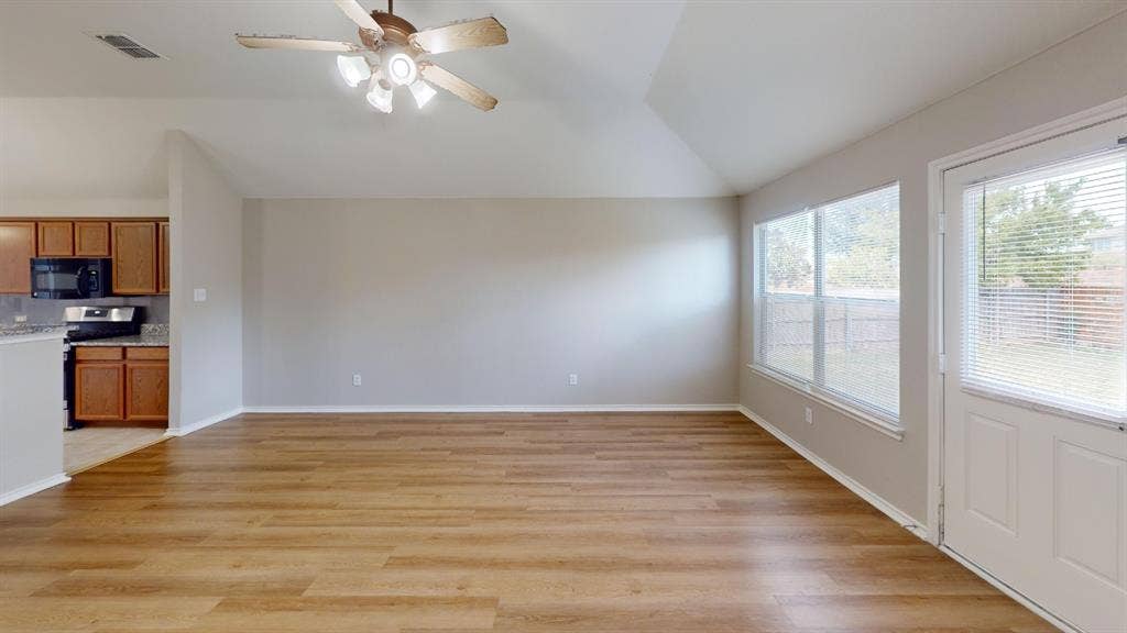 Vaulted ceiling featuring a central lighted ceiling fan, wood-finish flooring, dual windows, and an exterior door with integrated blinds