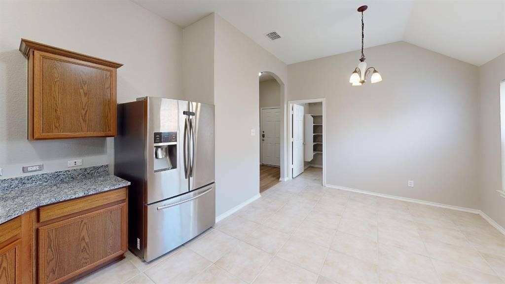 Vaulted ceiling featuring a pendant light fixture, stone-patterned countertops, stainless steel refrigerator, wood-finish cabinetry, and neutral tile flooring