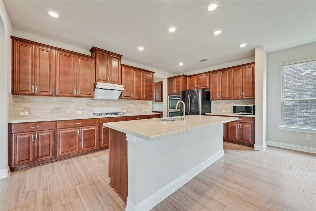 9829 Sharps Drive, Frisco, TX 75035 Kitchen featuring wood cabinetry, stone-finish countertops, a central island with an integrated sink, stone-finish tile backsplash, and wood-finish flooring