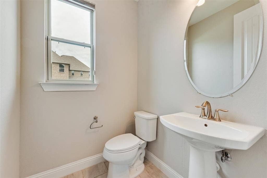 9829 Sharps Drive, Frisco, TX 75035 Pedestal sink with metallic faucet, oval wall-mounted mirror, white ceramic toilet, single-hung window, and light-tone tile flooring