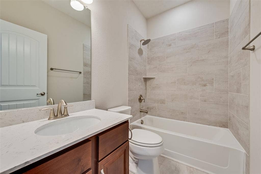 9829 Sharps Drive, Frisco, TX 75035 Bathroom featuring a wood-finish vanity with a stone-composite countertop, undermount sink, porcelain tile shower surround, and brushed metal fixtures