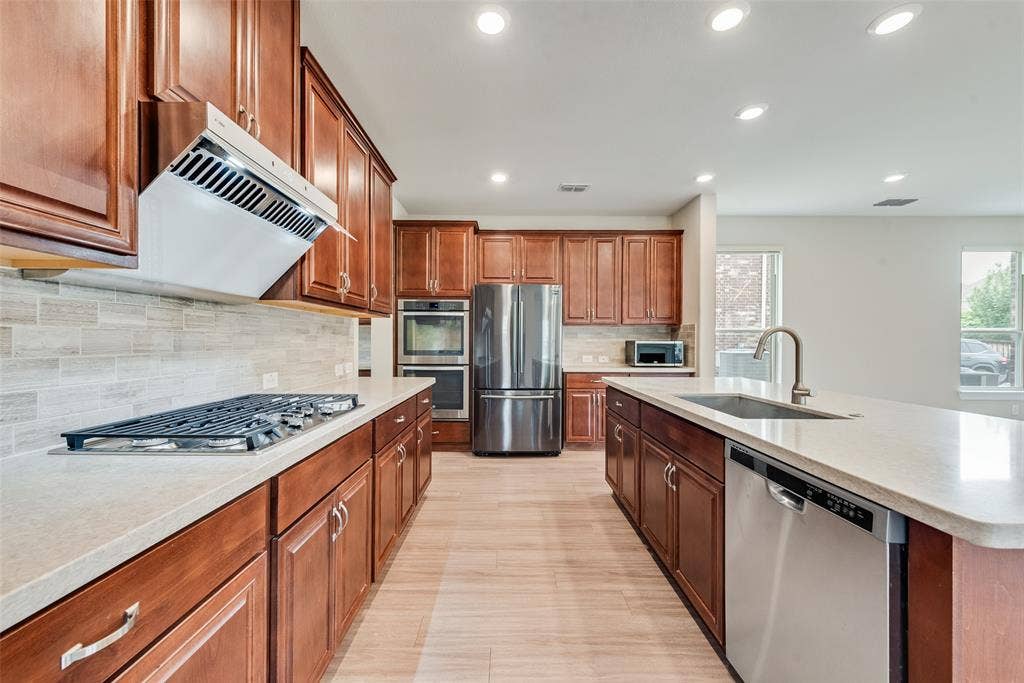 9829 Sharps Drive, Frisco, TX 75035 Kitchen featuring custom cabinetry, stone-finish countertops, stainless steel appliances including a built-in double oven and gas cooktop, tile backsplash, and a central island with a stainless steel sink