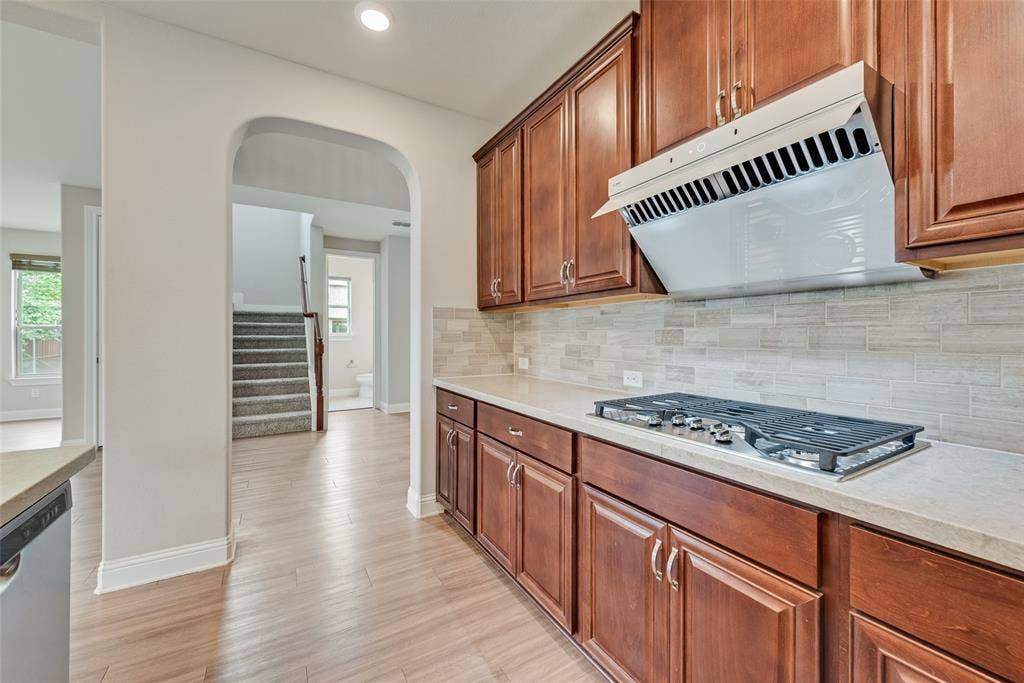 9829 Sharps Drive, Frisco, TX 75035 Kitchen featuring wood-finish cabinetry, stone-style countertops, linear tile backsplash, stainless steel gas cooktop, and a matching range hood