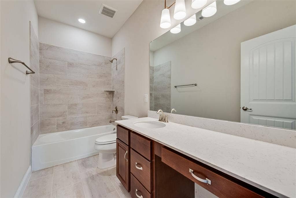9829 Sharps Drive, Frisco, TX 75035 Bathroom featuring a dual-drawer vanity with a stone-finish countertop, undermount sink, porcelain tile tub surround, and oversized wall-to-wall mirror with multi-bulb vanity lighting