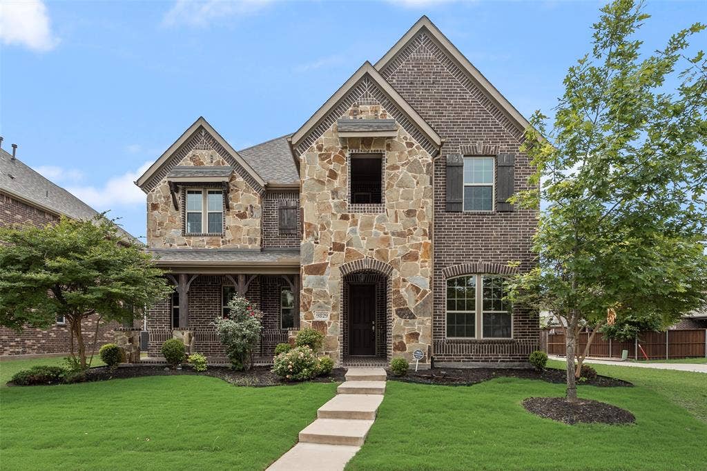 9829 Sharps Drive, Frisco, TX 75035 Two-story exterior featuring multi-gabled rooflines, a combination of dark brick and stone cladding, custom wood-finish shutters, and a covered front porch with timber beam detailing