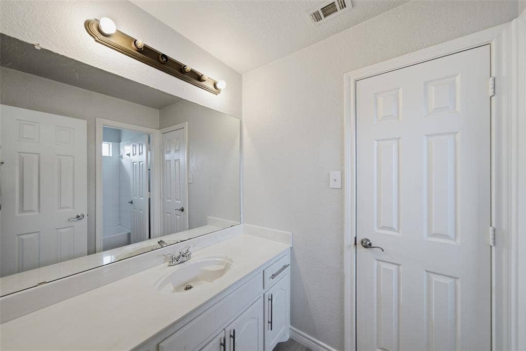 Bathroom vanity featuring a solid-surface countertop, an integrated sink, white cabinetry with silver-tone hardware, and a large wall-mounted mirror with an attached multi-bulb light fixture