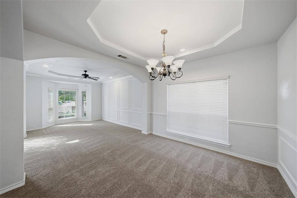 Tray ceiling featuring a decorative chandelier, wainscoting, arched architectural detail, neutral-toned carpeting, and a bay window