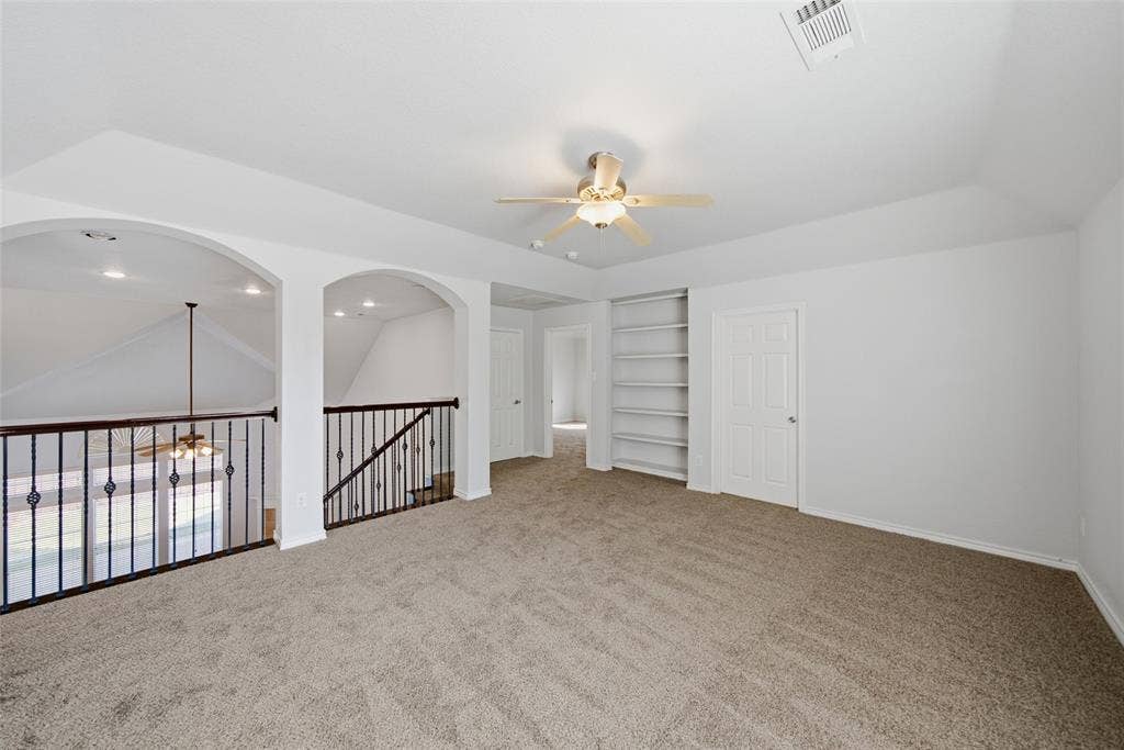 Open area featuring tray ceilings, carpeted flooring, built-in shelving, a ceiling fan, and decorative arched architectural details with metal railings