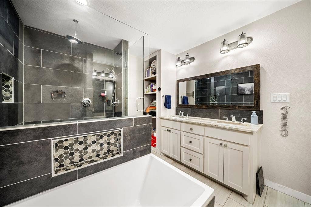 205 Newman Road, Ennis, TX 75119 Bathroom featuring a glass-enclosed shower with large-format tile, a built-in recessed niche with hexagonal mosaic accents, a double-sink vanity with a light-toned stone countertop, and built-in floor-to-ceiling shelving