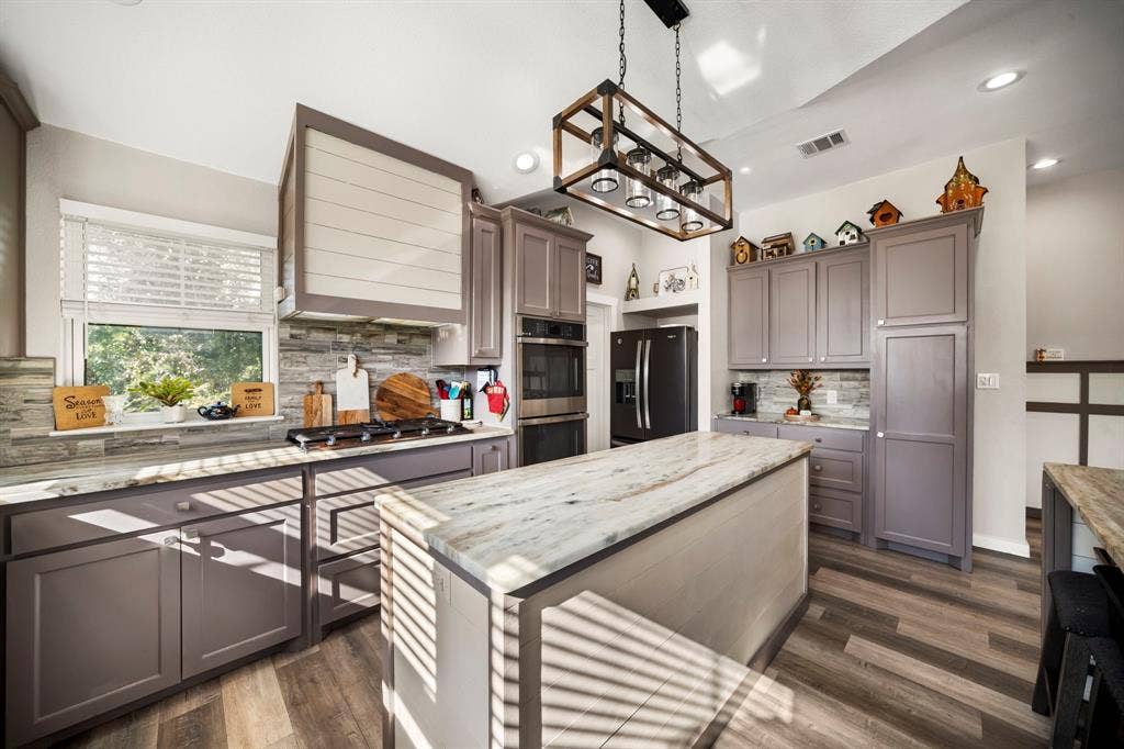 205 Newman Road, Ennis, TX 75119 Kitchen featuring a central island with a stone countertop, grey shaker-style cabinetry, horizontal-plank range hood surround, stone-tile backsplash, and rectangular lantern-style pendant lighting