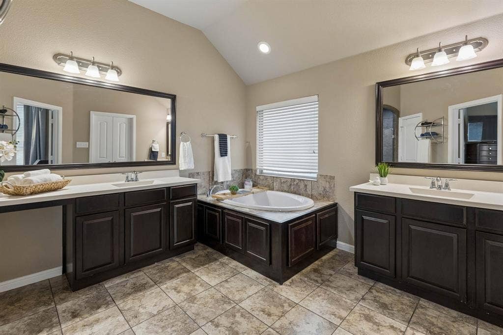 The primary bath features dual vanities, stained wood cabinets, ample storage, tile flooring, a relaxing garden tub, recessed lighting, framed mirrors, and a spacious walk-in closet.
