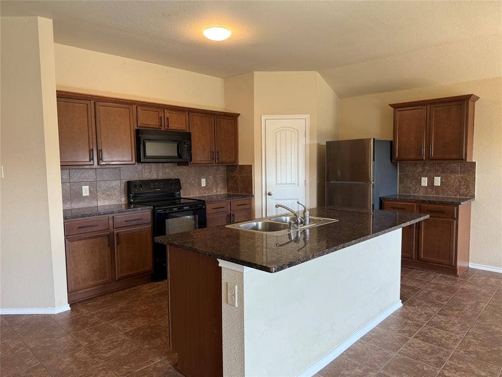 212 Belford Street S, Anna, TX 75409 Kitchen featuring wood cabinetry, a central island with a dual-basin sink, stone-finish countertops, tile backsplash, and tile flooring