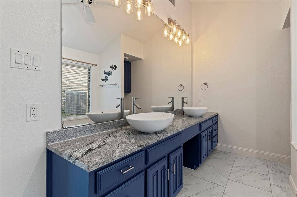 4128 Woodenrail Lane, Irving, TX 75061 Dual vanity featuring navy blue cabinetry, stone-finish countertops, white vessel sinks, and tiered glass-shade light fixtures, complemented by large-format floor tile