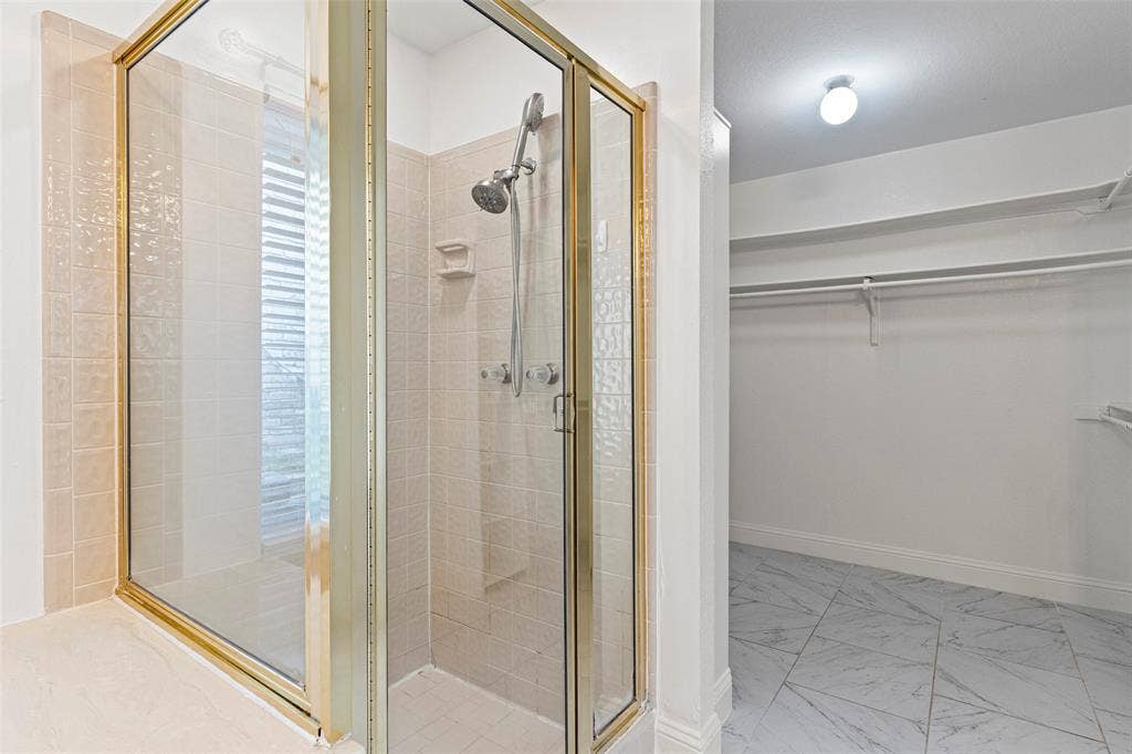 4128 Woodenrail Lane, Irving, TX 75061 Walk-in shower featuring a metallic frame, hand-held showerhead, and tiled surround, paired with a storage area finished with marble-patterned tile flooring and built-in shelving