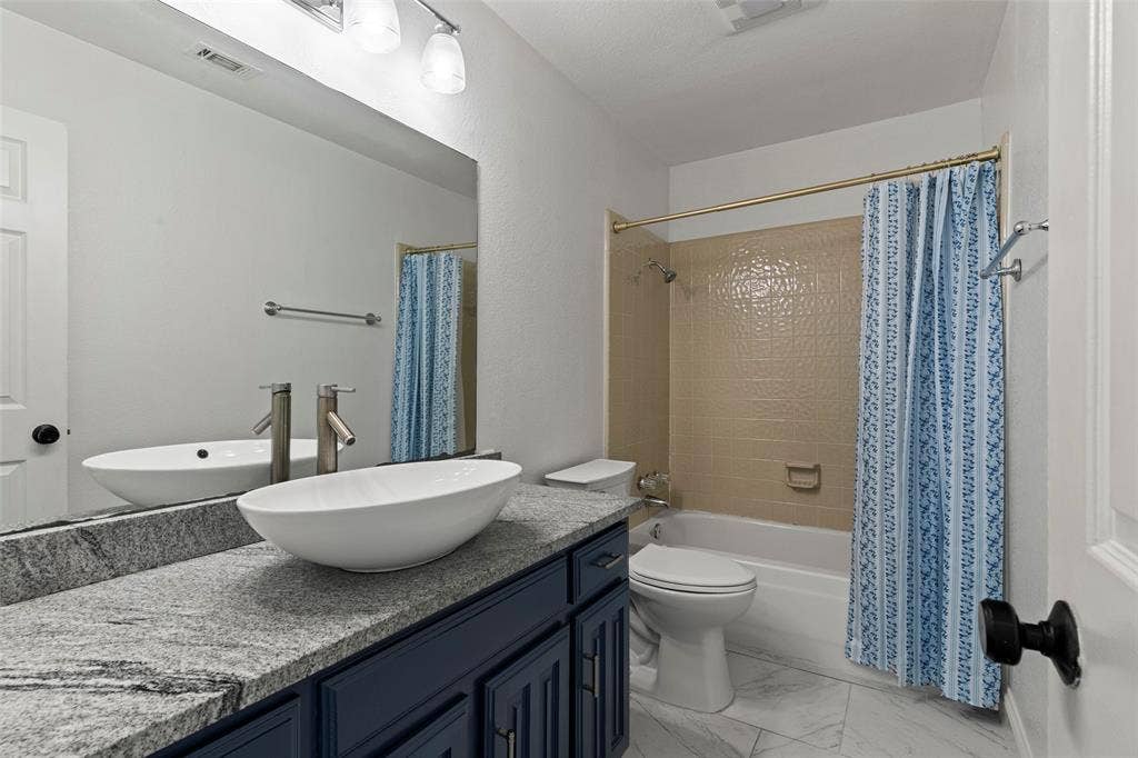 4128 Woodenrail Lane, Irving, TX 75061 Bathroom featuring a vanity with dark-finish cabinetry, stone countertop, and an oval vessel sink, complemented by stone-patterned tile flooring and a tiled shower surround