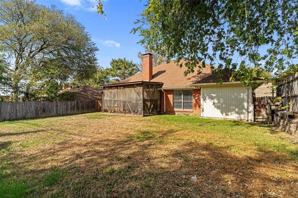 4128 Woodenrail Lane, Irving, TX 75061 Rear exterior featuring a brick chimney, screened-in porch, composition shingle roof, wood privacy fencing, and a retaining wall