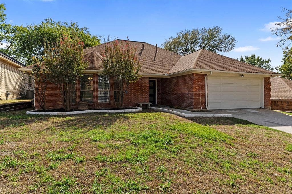 4128 Woodenrail Lane, Irving, TX 75061 Single-story brick exterior featuring a multi-gabled roofline, an attached two-car garage, and stone-bordered landscaping beds