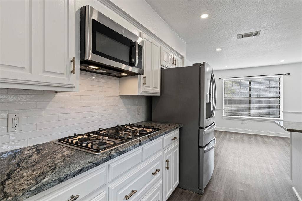 4128 Woodenrail Lane, Irving, TX 75061 Kitchen featuring white shaker-style cabinetry, granite countertops, a subway tile backsplash, a built-in stainless steel microwave, and wood-finish flooring