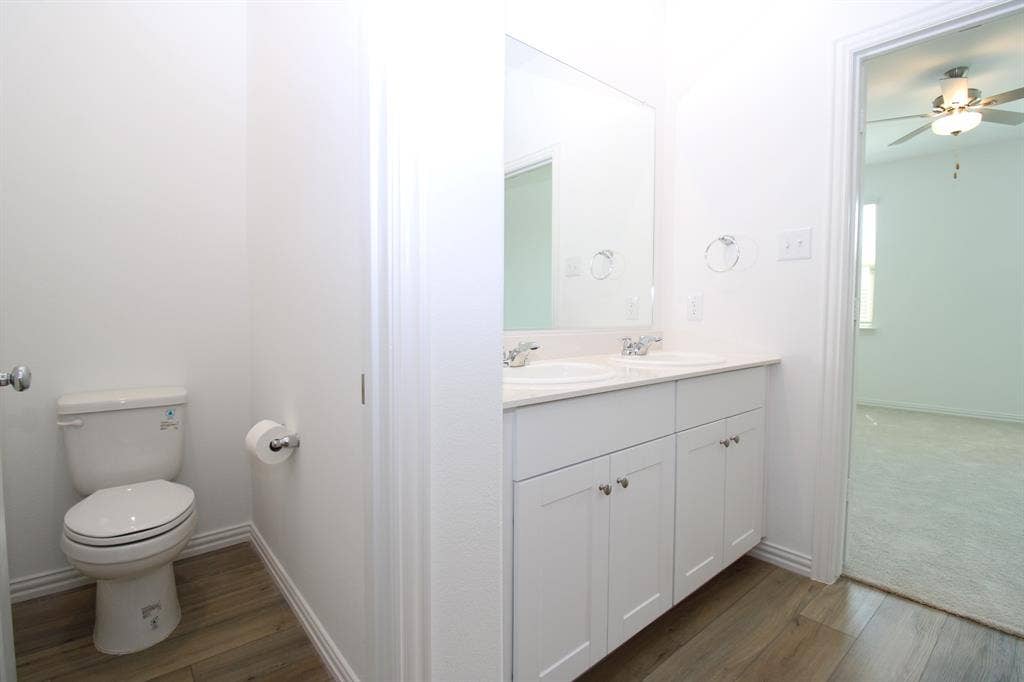 Dual-sink vanity with white shaker-style cabinetry, light-toned countertop, wood-finish flooring, and wall-mounted mirror