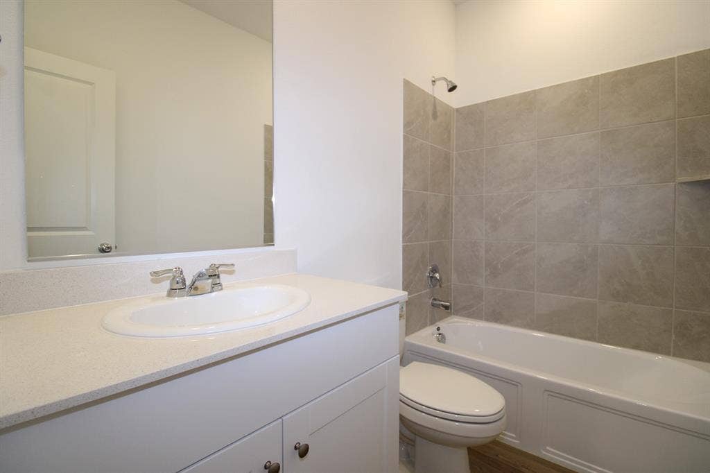 Bathroom featuring a vanity with a stone-surface countertop, an integrated oval sink, a wall-mounted mirror, a porcelain tub with a tiled surround, and a showerhead fixture