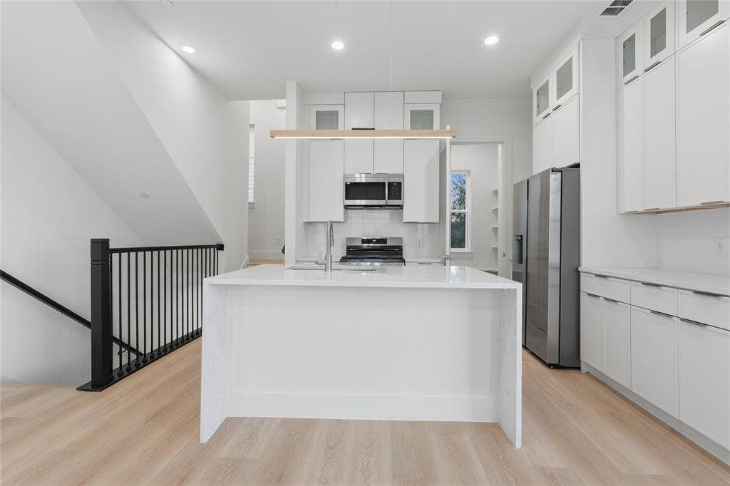 4319 San Jacinto Street, #101, Dallas, TX 75204 Kitchen featuring an expansive waterfall-edge island, white shaker-style cabinetry with glass-front uppers, stainless steel appliances, and wood-finish flooring