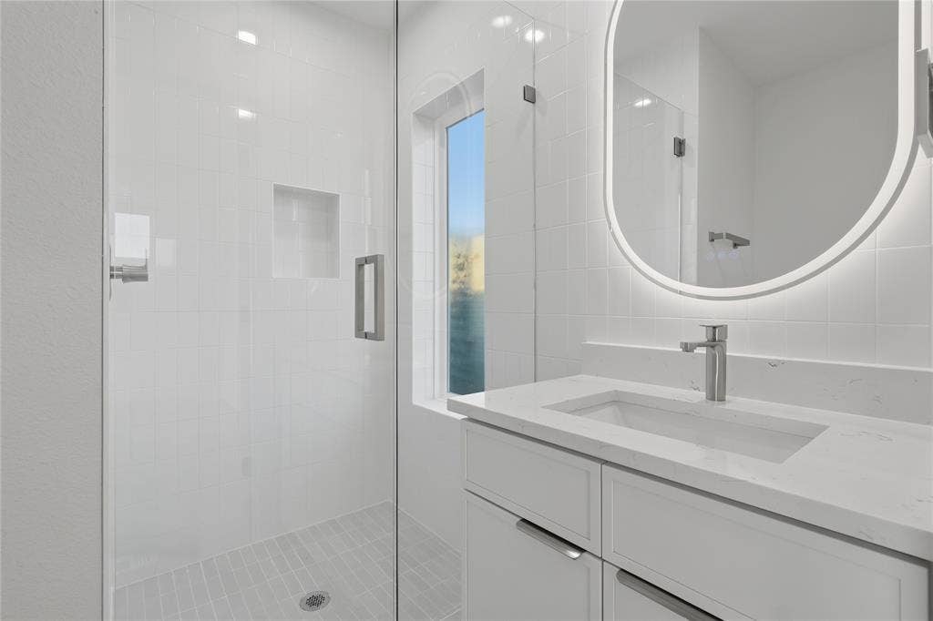 4319 San Jacinto Street, #101, Dallas, TX 75204 Bathroom featuring a white vanity with a stone countertop, an integrated rectangular sink, a frameless glass shower enclosure with a recessed tile niche, and an oval backlit mirror