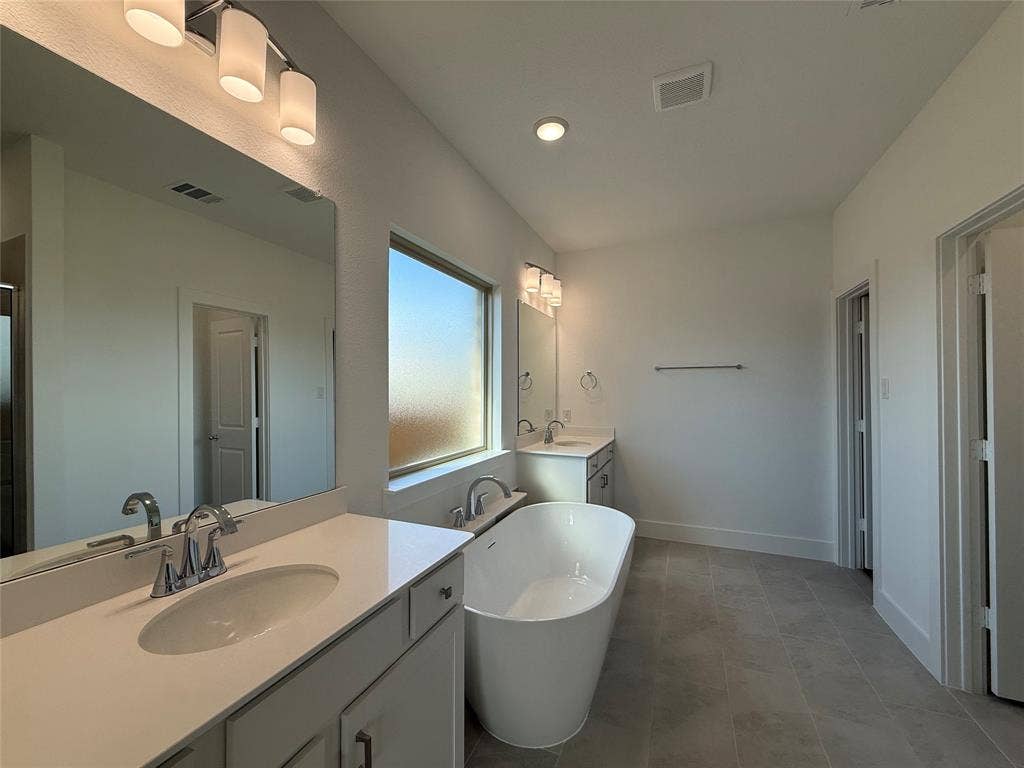 Bathroom featuring a freestanding oval soaking tub, white cabinetry with stone-finish countertops, dual-sink configuration, large wall-mounted mirrors, and tiled flooring