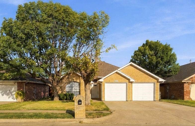 Single-story brick exterior with a dual-car garage, concrete driveway, and established landscape featuring mature trees
