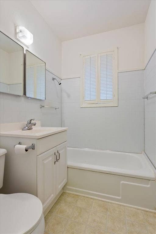 3907 Rogers Hill Road, Waco, TX 76705 Bathroom featuring a vanity with a white cabinet and integrated sink, a tiled tub surround, a window with louvered shutters, wall-mounted lighting, and rectangular wall mirrors