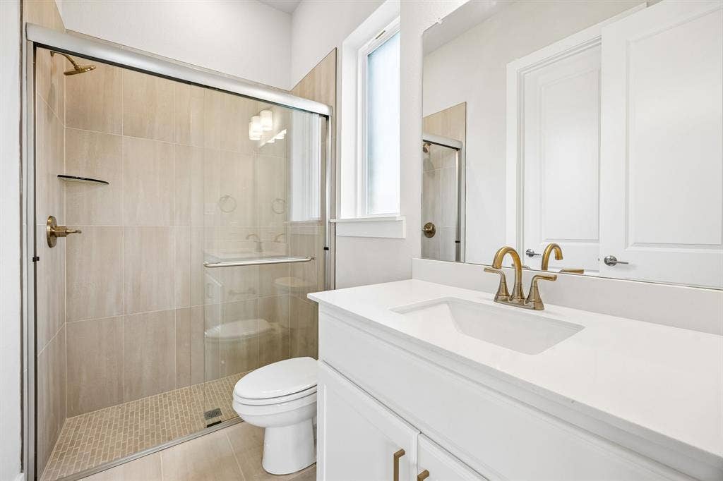 108 Swan Lake Drive, Waxahachie, TX 75165 Bathroom featuring a white vanity with a rectangular sink, stone-finish countertop, brass-tone hardware, glass-enclosed shower with tiled walls and flooring, and a large wall mirror