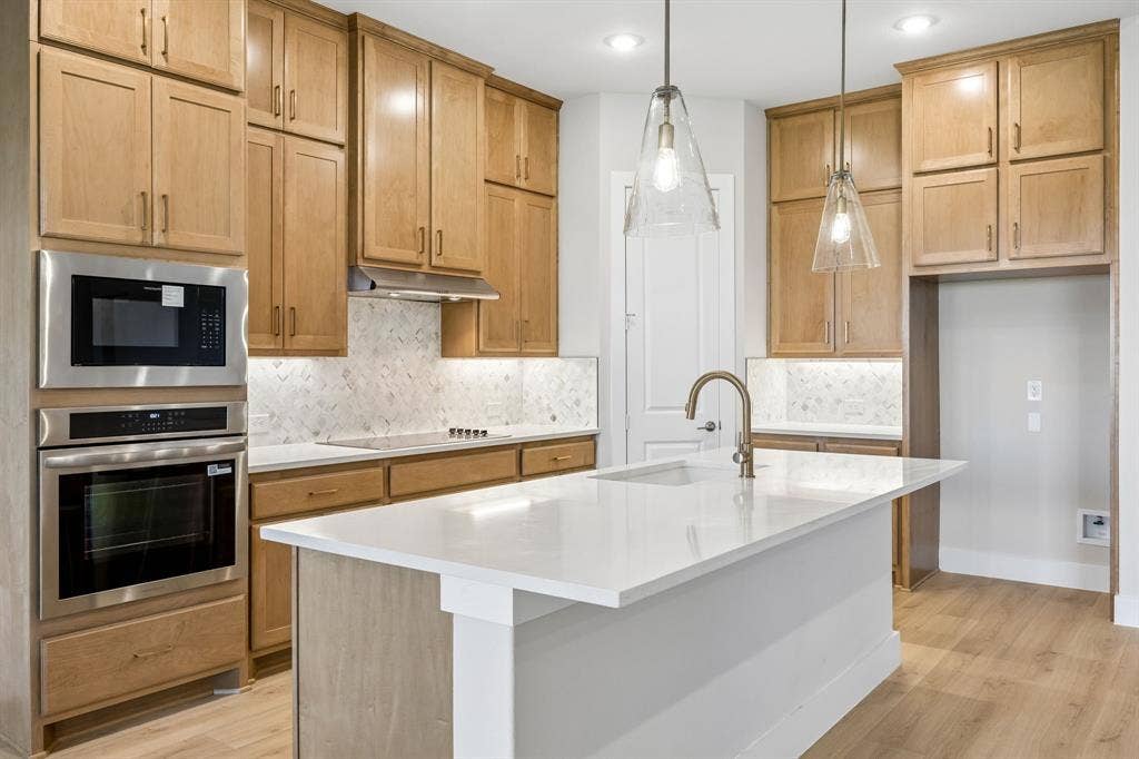 108 Swan Lake Drive, Waxahachie, TX 75165 Kitchen featuring light wood-finish cabinetry, white stone countertops, herringbone-pattern tile backsplash, stainless steel wall-mounted oven and microwave, and a central island with pendant lighting