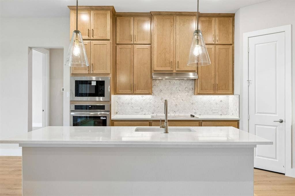 108 Swan Lake Drive, Waxahachie, TX 75165 Kitchen featuring custom-height wood cabinetry, quartz countertop kitchen island with undermount sink, chevron-pattern tile backsplash, stainless steel built-in double wall oven and microwave, and glass pendant lighting