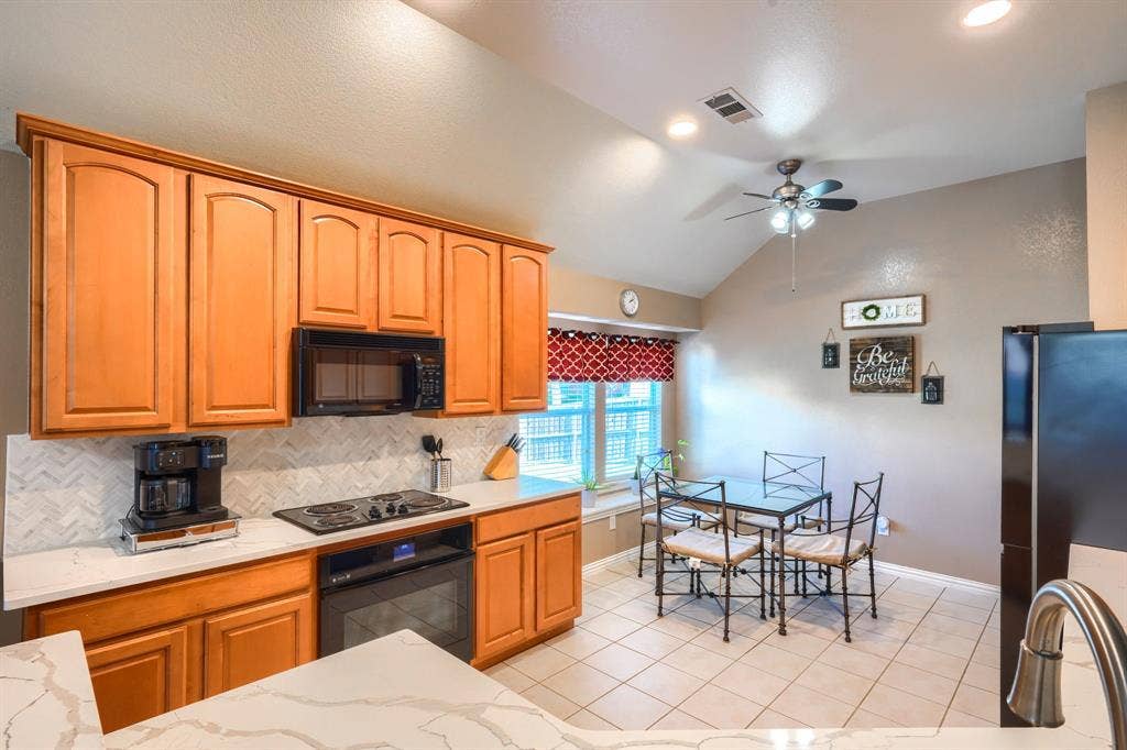 Kitchen featuring natural wood-finish cabinetry, white stone-composite countertops, a herringbone-patterned tile backsplash, a vaulted ceiling, and tile flooring