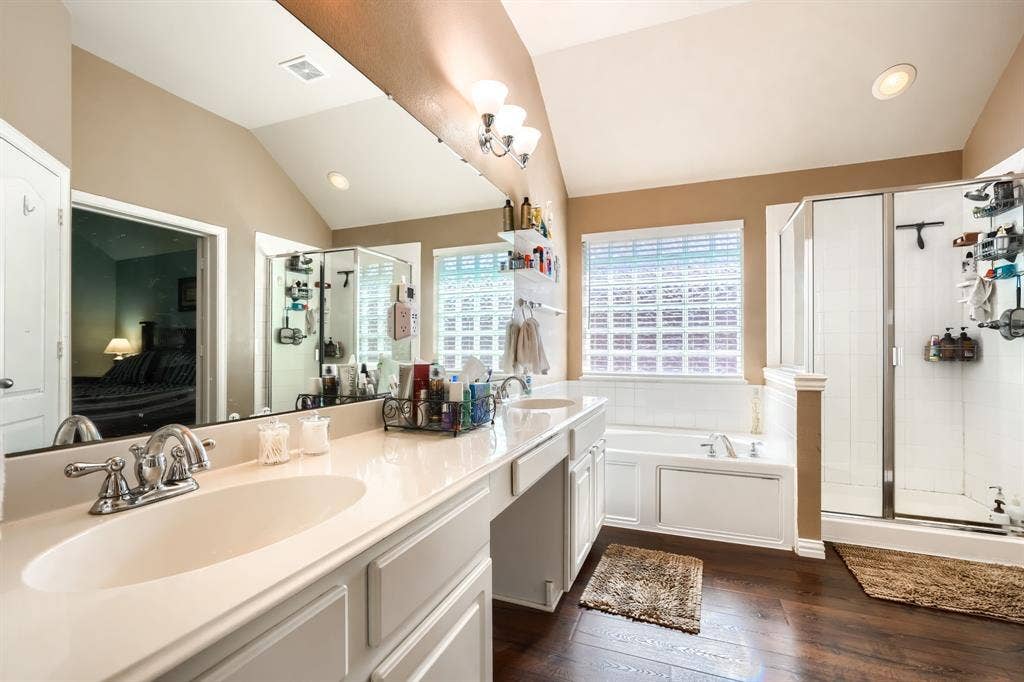 Bathroom featuring vaulted ceilings, dual-sink vanity with solid surface countertop, soaking tub, glass-enclosed shower, and wood-finish flooring