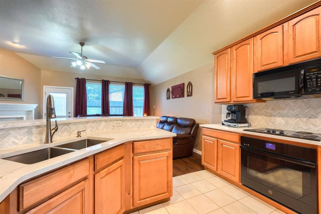 Kitchen featuring wood cabinetry, stone-finish countertops, herringbone-pattern tile backsplash, stainless steel dual-basin sink with pull-down faucet, and built-in black oven and microwave