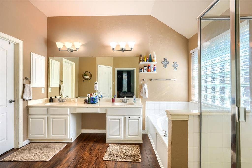 Dual-sink vanity with white cabinetry and a solid-surface countertop, full-width vanity mirror, glass-enclosed shower, built-in soaking tub with tile surround, and wood-finish flooring