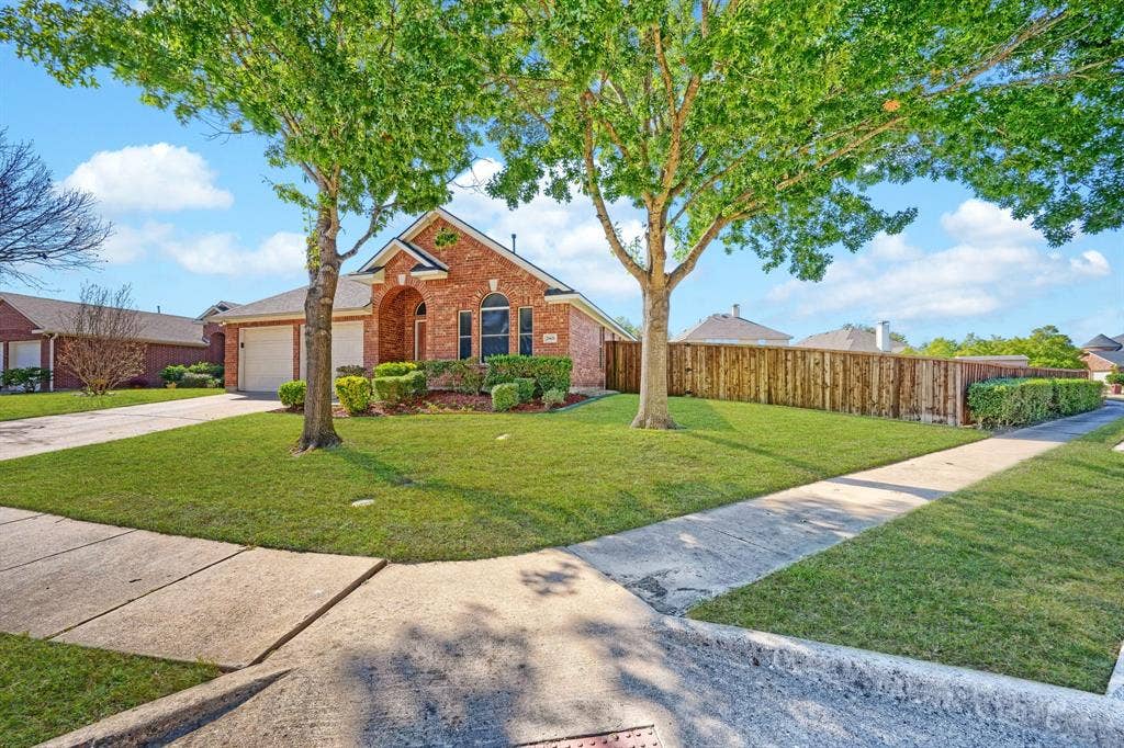Brick exterior residence featuring an arched entryway, gabled roofline, concrete driveway, and a fenced yard with mature trees