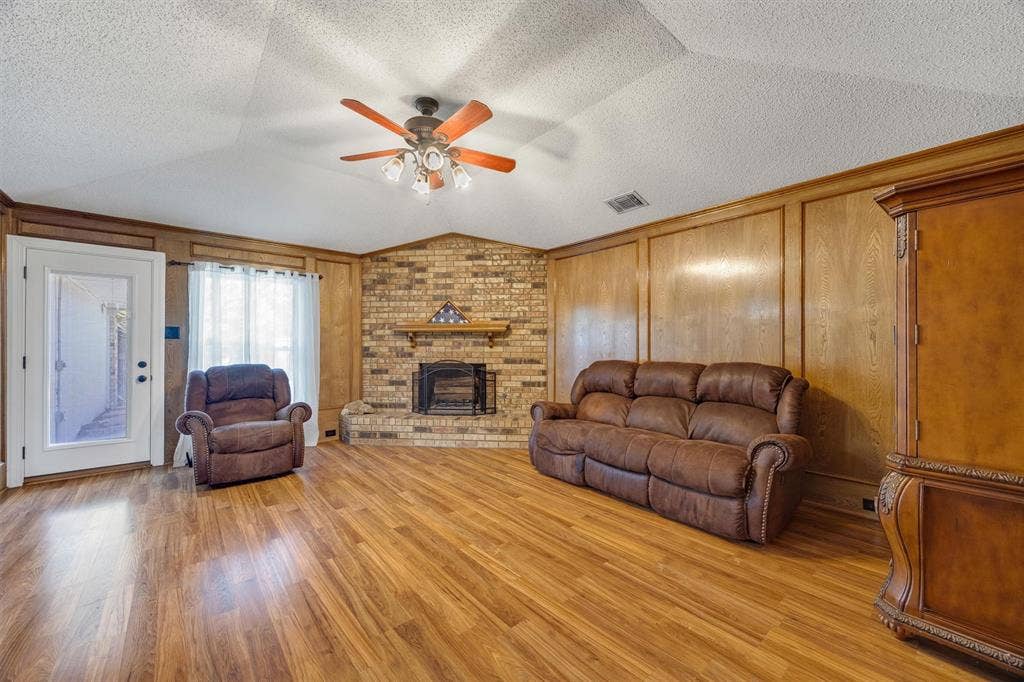 1025 Brownfield Drive, Mesquite, TX 75150 Vaulted ceiling with a central multi-blade ceiling fan, full-height wood paneling, wood-finish flooring, and a brick fireplace featuring a wood mantel