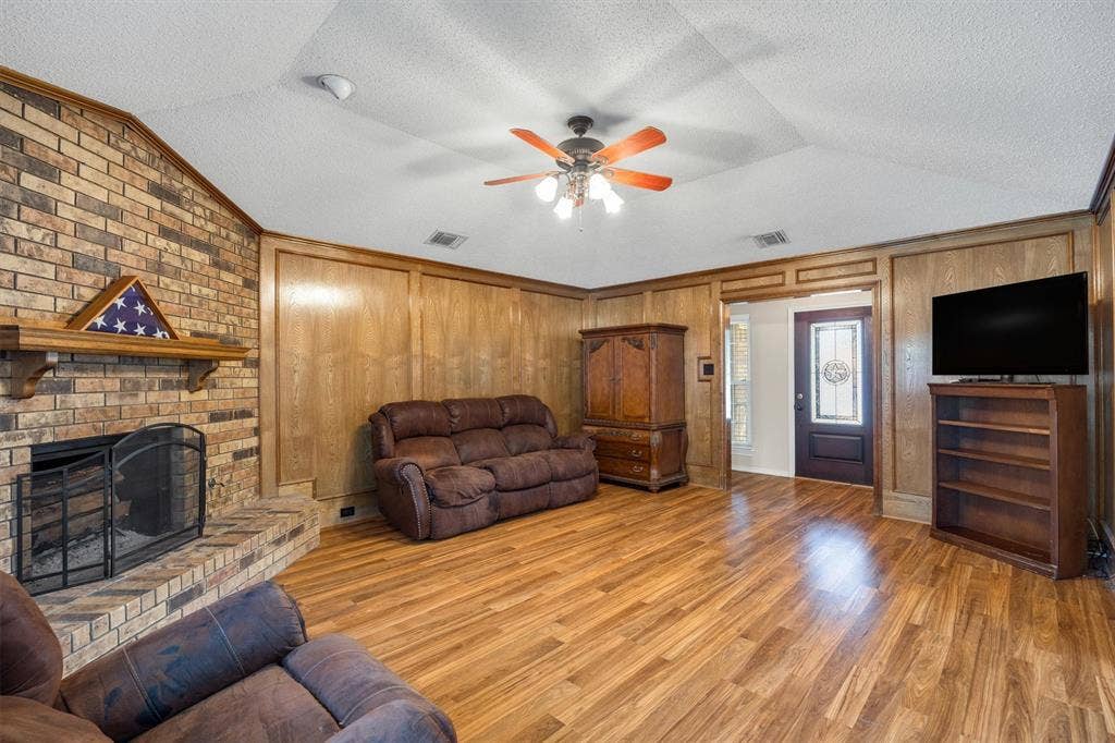 1025 Brownfield Drive, Mesquite, TX 75150 Vaulted ceiling with a central multi-blade ceiling fan, floor-to-ceiling brick fireplace with a wood mantel, wood-finish paneling, and wood-finish flooring