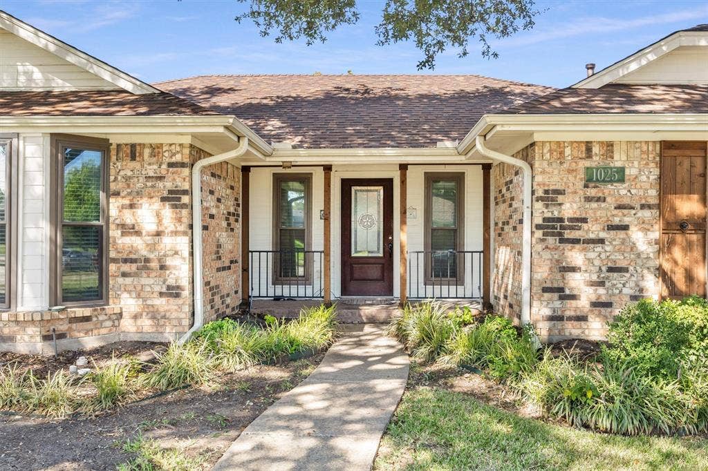 1025 Brownfield Drive, Mesquite, TX 75150 Brick exterior facade featuring a covered front porch, dark-stained wooden support posts, black metal railings, and a decorative glass-panel front door
