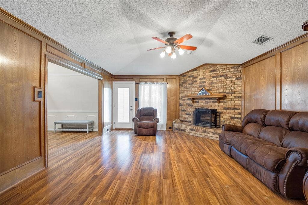 1025 Brownfield Drive, Mesquite, TX 75150 Living area featuring wood-finish flooring, full-height wood wall paneling, a vaulted ceiling with a central fan, and a masonry brick fireplace with a wood mantel