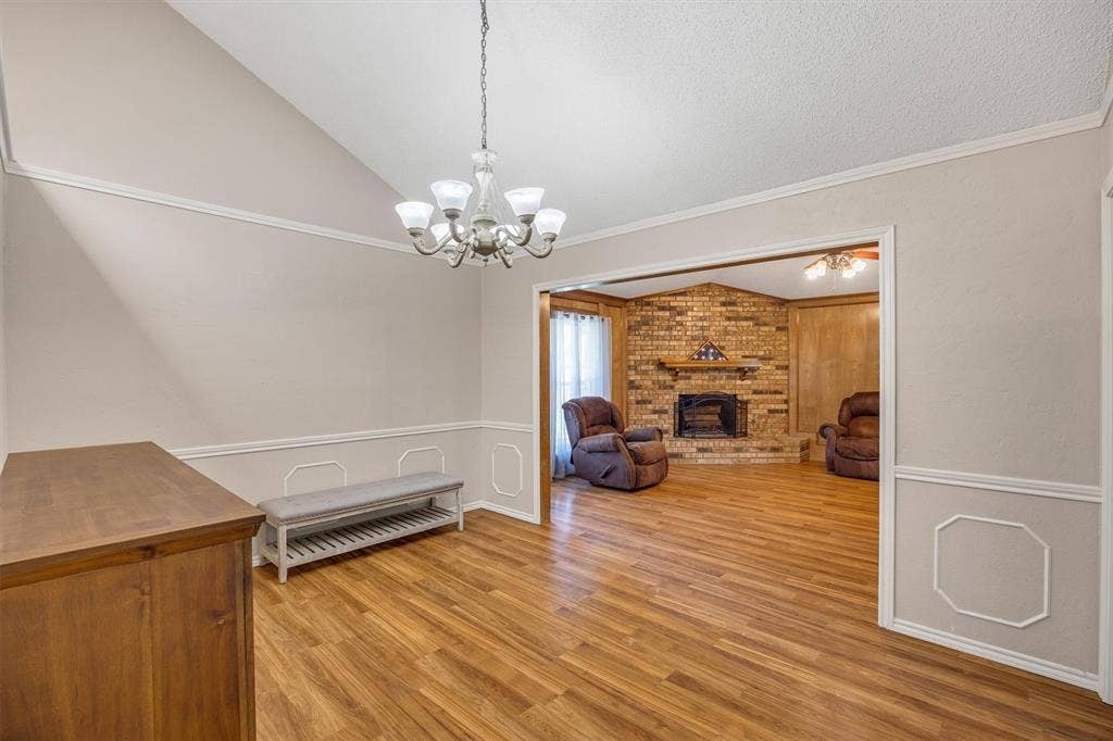 1025 Brownfield Drive, Mesquite, TX 75150 Vaulted ceiling with a multi-arm chandelier, wood-finish flooring, decorative wall molding, and a brick fireplace featuring a wood mantel
