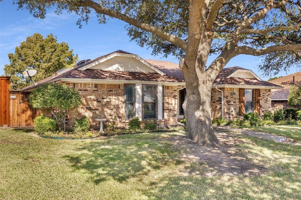 1025 Brownfield Drive, Mesquite, TX 75150 Single-story brick exterior featuring a hip roof, multi-pane windows, wood privacy fencing, and a mature tree