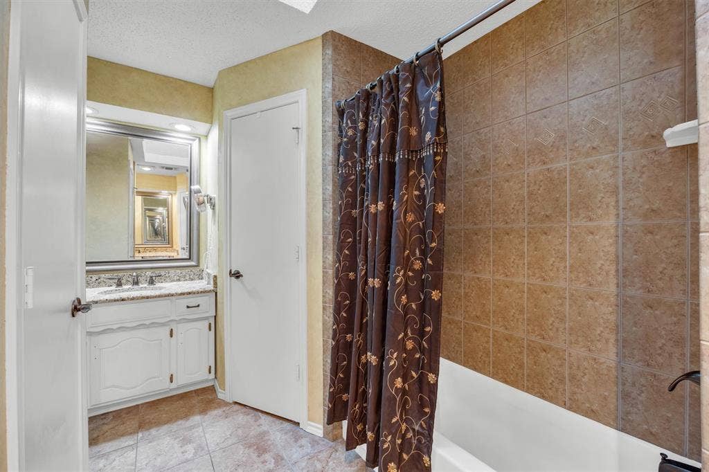 1025 Brownfield Drive, Mesquite, TX 75150 Bathroom featuring a stone-patterned vanity countertop, white cabinetry with integrated hardware, a framed wall mirror, tiled flooring, and a bathtub enclosure with decorative wall tile