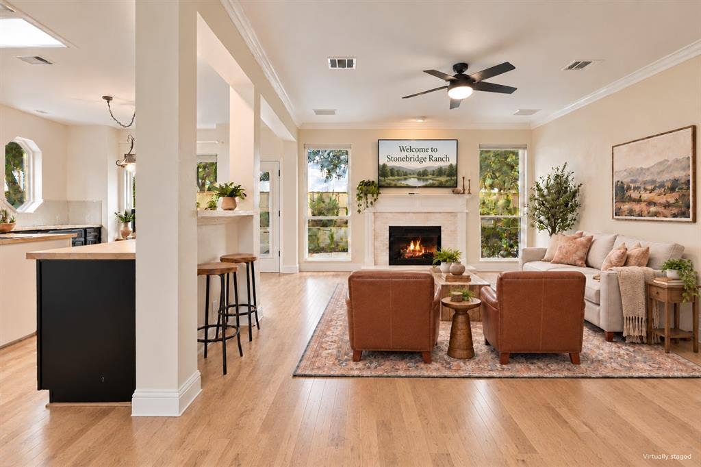 511 Clover Leaf Lane, McKinney, TX 75072 Open-concept room featuring light wood-finish flooring, a neutral-toned fireplace surround with a mantle, crown molding, a ceiling fan, and large windows