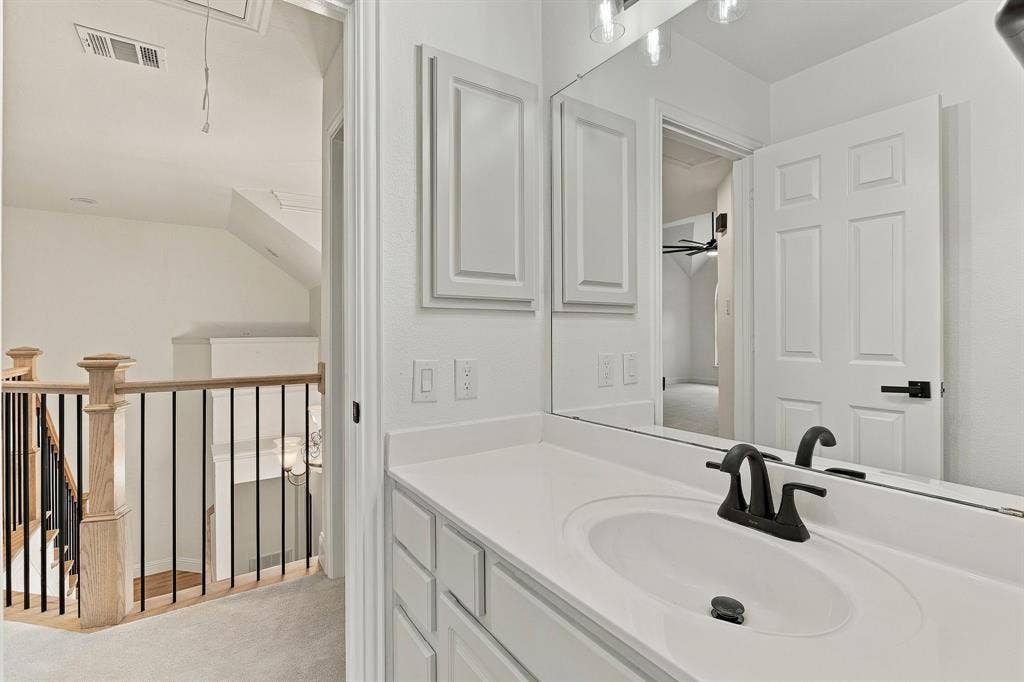 511 Clover Leaf Lane, McKinney, TX 75072 Vanity featuring a white countertop with an integrated oval basin and matte black faucet, full-width mirror, recessed cabinetry, and a staircase with a wooden banister and black metal balusters