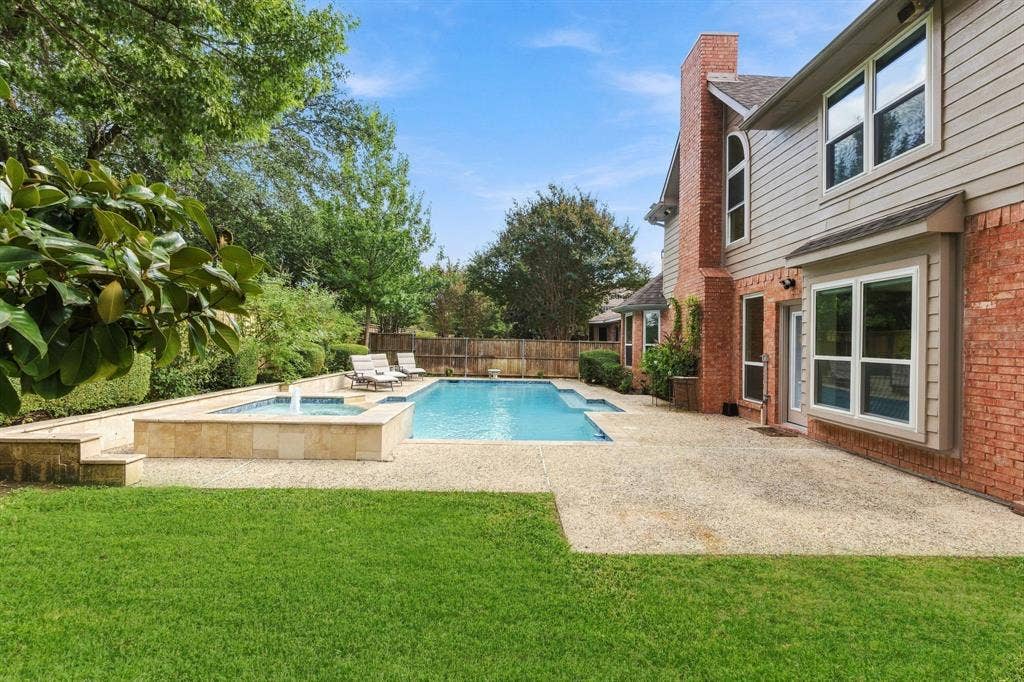 511 Clover Leaf Lane, McKinney, TX 75072 Rear exterior featuring a brick and siding facade, an in-ground swimming pool with an integrated spa, a tiered stone retaining wall, and a sprawling concrete patio