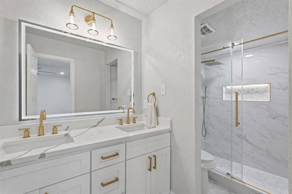 5514 Round Rock Road, Garland, TX 75044 Dual-sink vanity with stone-surfaced countertop, integrated LED-backlit mirror, brass-finish hardware and fixtures, and a walk-in shower featuring a glass enclosure with a tiled built-in niche