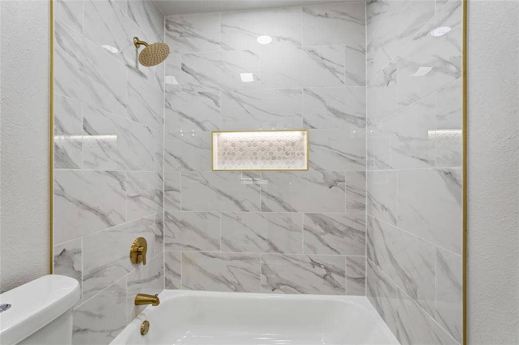 5514 Round Rock Road, Garland, TX 75044 Bathtub with surround featuring large-format stone-patterned tile, gold-tone metal trim, integrated recessed niche with mosaic tiling, and gold-tone faucet and shower fixtures