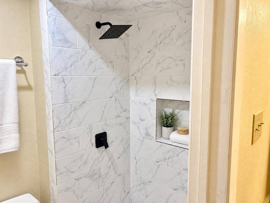 618 Pyramid Drive, Garland, TX 75040 Shower enclosure featuring large-format veined tile, matte black rainfall showerhead, matching single-handle valve, and a built-in recessed storage niche